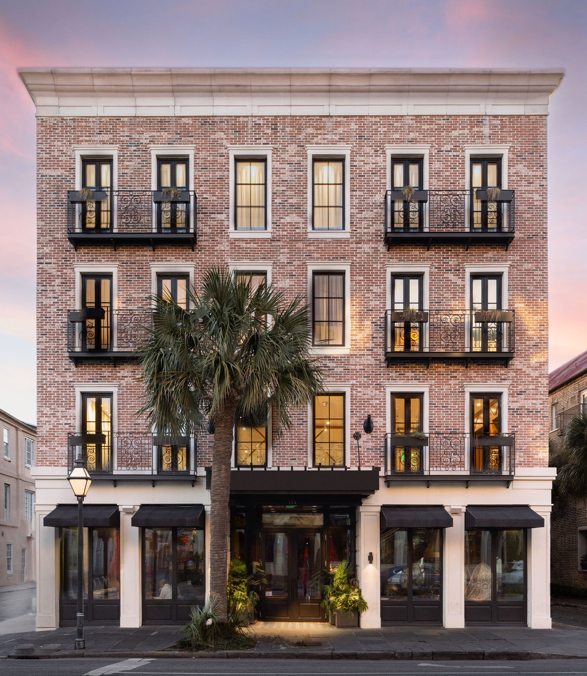 A multi-story brick building with large windows and small balconies on the second and third floors. The building has a black awning at the entrance and is flanked by a street lamp and a palm tree in front. Lights are on inside, and the sky is pink an