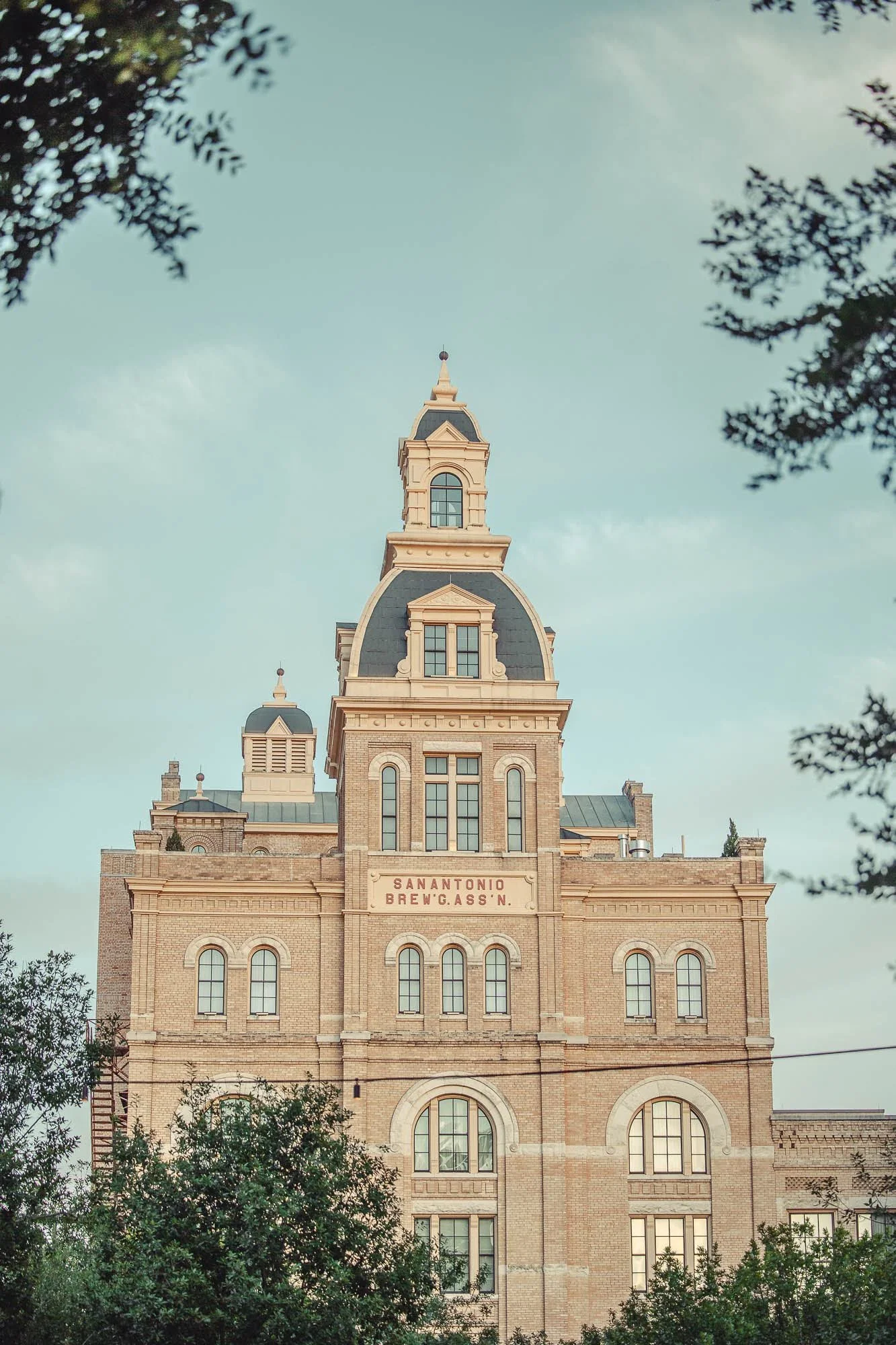 Historic brick building with a tall central clock tower, trees partially obscuring the lower part of the building, and a sign that reads 'San Antonio Brewing Ass'n' on the facade.