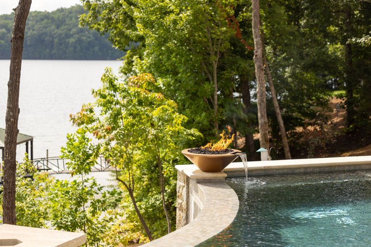 A hot tub outdoors near a lake with trees, featuring a fire bowl on the edge with flames and water flowing from the bowl into the hot tub.