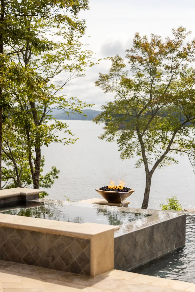 An outdoor setting with a pool overlooking a lake, trees with green leaves, and a fire feature on the pool's edge.