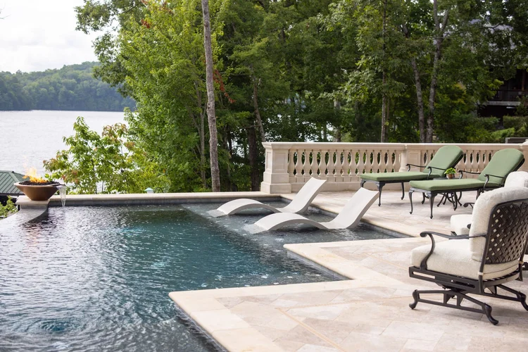 A backyard infinity pool overlooking a lake, with lounge chairs, patio furniture, and a fire bowl on the deck surrounded by trees.