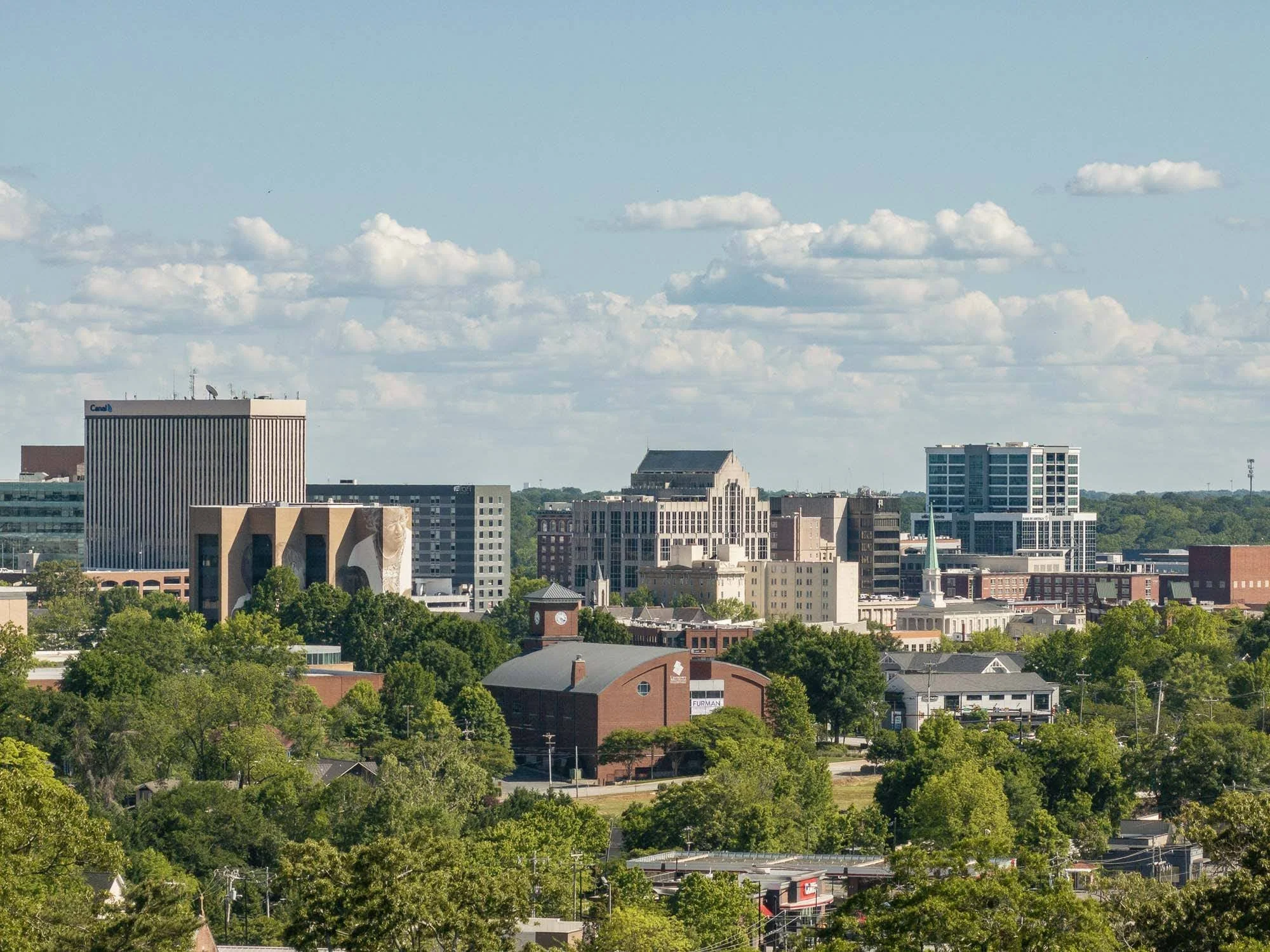 Urban cityscape with various modern and historic buildings, surrounded by green trees, under a partly cloudy sky.