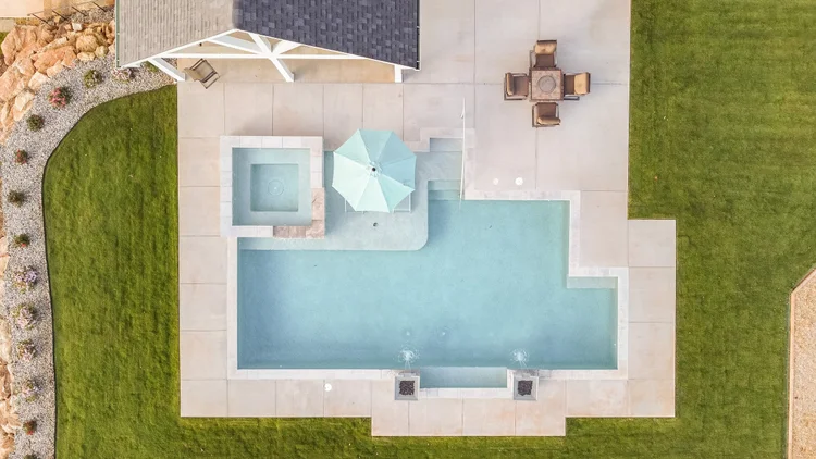 An aerial view of a backyard with a swimming pool, a small spa, a patio area with a table and chairs, a grassy lawn, and a decorative rock and plant border.