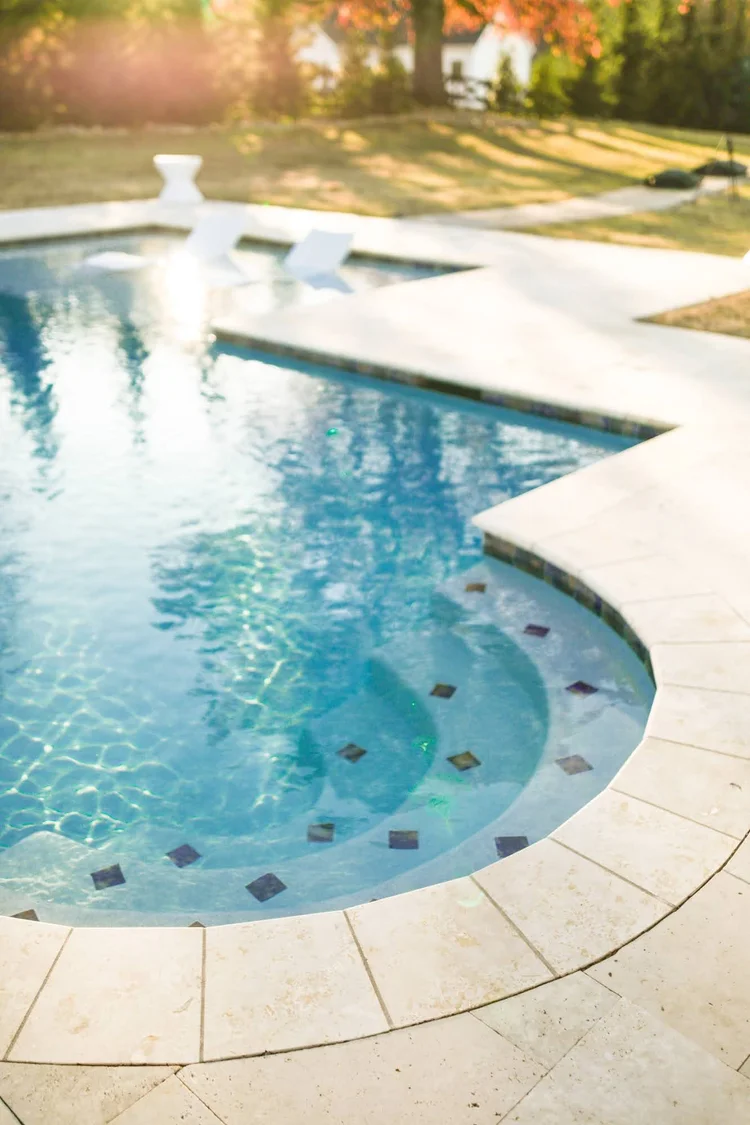 Residential backyard swimming pool with two white lounge chairs, surrounded by a stone deck and fenced yard with trees and autumn foliage in the background.