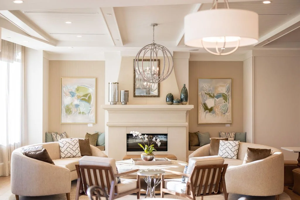 A bright, modern living room with beige sofas, a central coffee table with a plant, wall art, and a white fireplace with decorative vases on top. Large windows with curtains allow natural light.