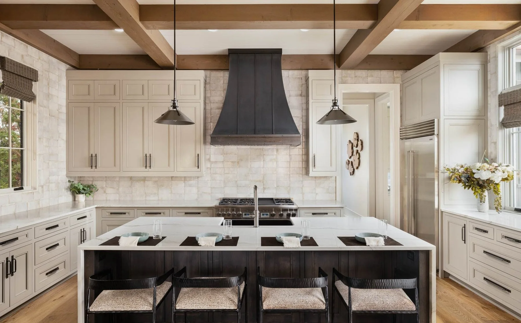 A modern kitchen featuring white cabinets, a central island with black paneling, a black range hood, stainless steel appliances, and large windows with blinds.