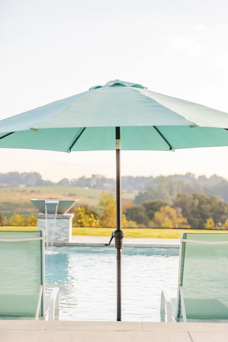 A swimming pool area with a large blue umbrella, two poolside chairs, and a scenic rural landscape with rolling hills and trees in the background.