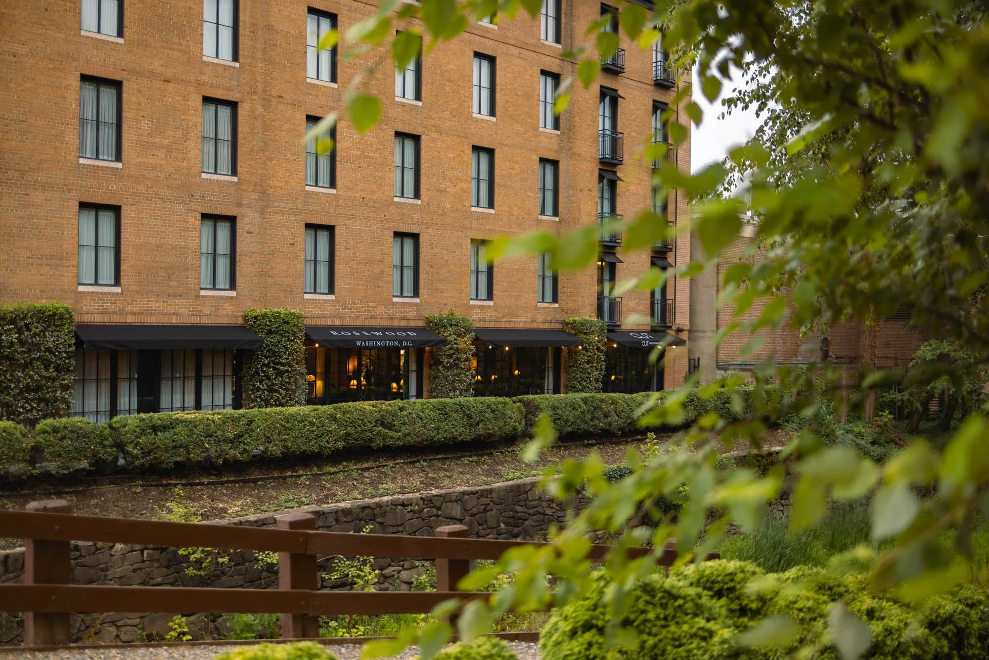A brick building with multiple floors and large windows, with some of the windows having black balconies. The ground floor appears to be a restaurant or cafe with black awnings and the words 'Washington, D.C.' visible on one. There are landscaped bus