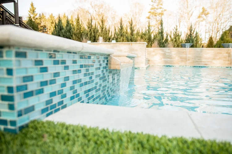 Close-up view of the edge of a swimming pool with blue mosaic tiles, water flowing from a spout into the pool, and a brick wall with planters in the background.