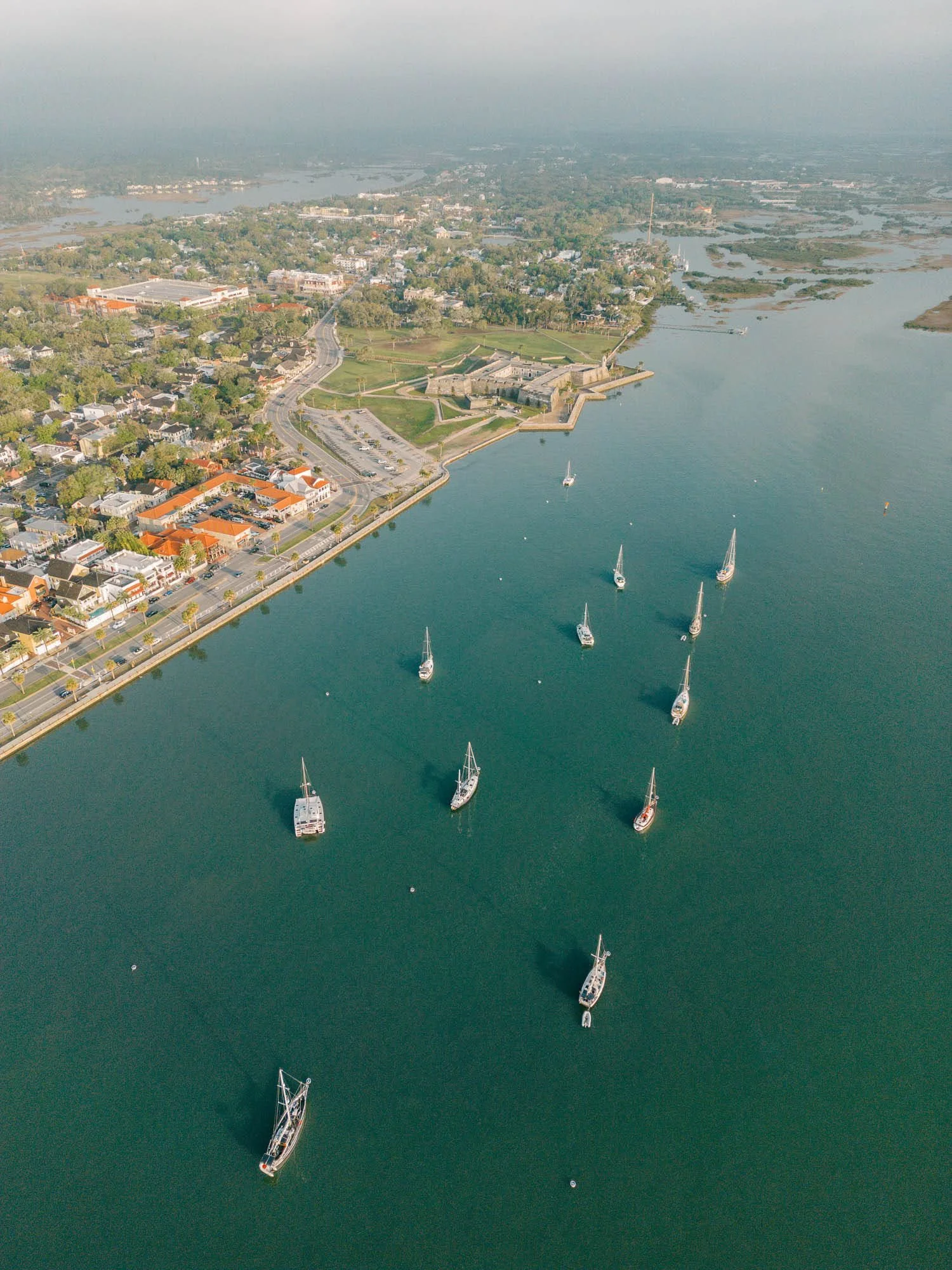 An aerial view of a coastal town with boats docked in the water, historic fortress walls along the shoreline, residential buildings, and green parks.