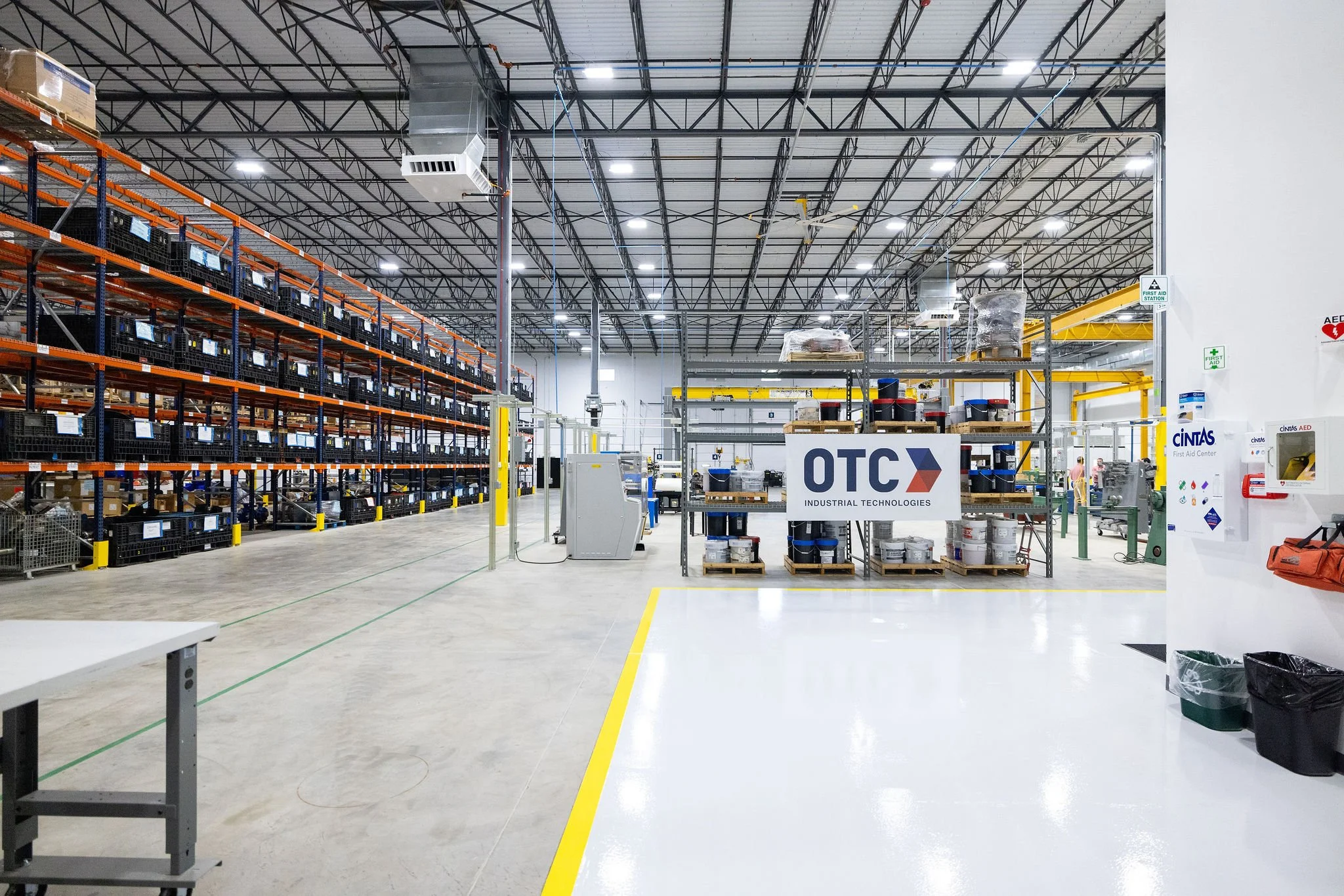 Interior of a large warehouse or industrial store with metal shelves filled with black crates and some boxed items. There are yellow safety barriers, a white wall on the right with signs for first aid and AED, and a high ceiling with metal beams and 