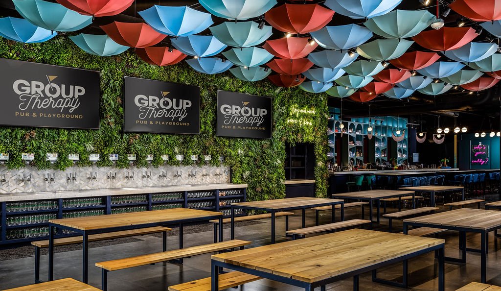 Interior of a bar or pub with wooden tables and benches, decorated with colorful umbrellas hanging from the ceiling, green wall with TVs displaying "Group Therapy" and neon signs.