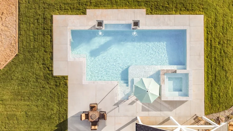 Aerial view of a backyard with a large rectangular swimming pool, small hot tub, patio furniture, and a green umbrella, surrounded by a grassy yard.