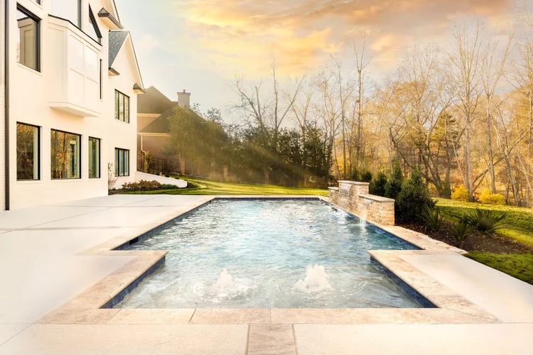 A backyard swimming pool with water feature next to a white house, surrounded by a grassy lawn and trees during sunset.