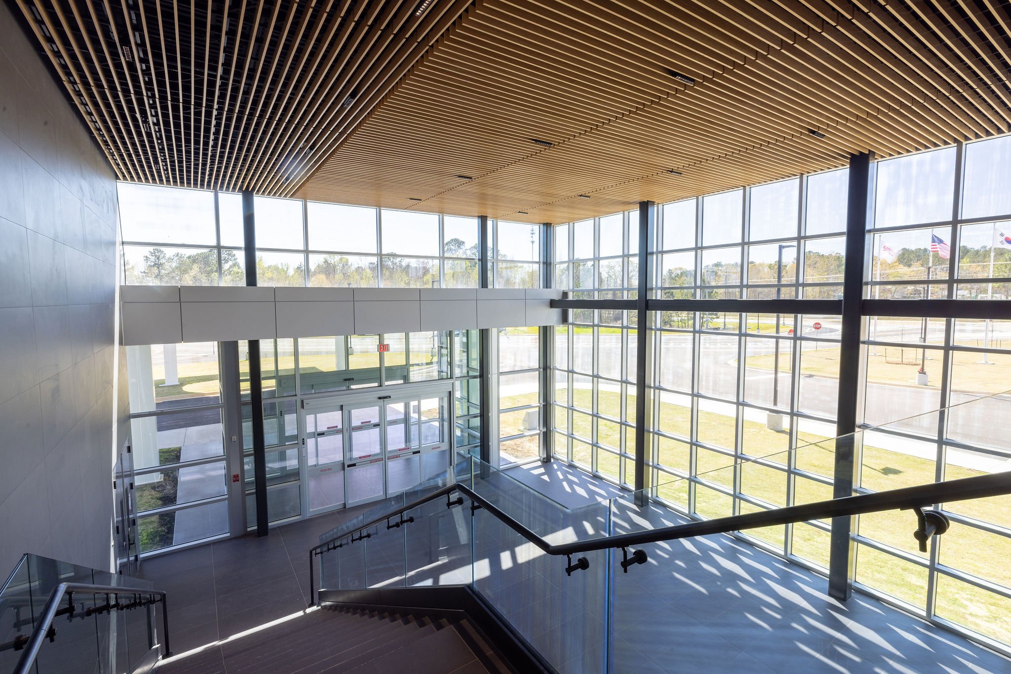 Interior of a modern building with large glass windows, a sloped staircase with metal railings, a wooden ceiling with evenly spaced slats, and outside view of flags and trees.