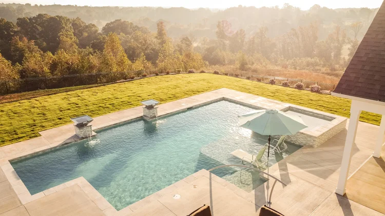 A backyard swimming pool with a hot tub, surrounded by a concrete deck, on a sunny day with a grassy yard and trees in the background.