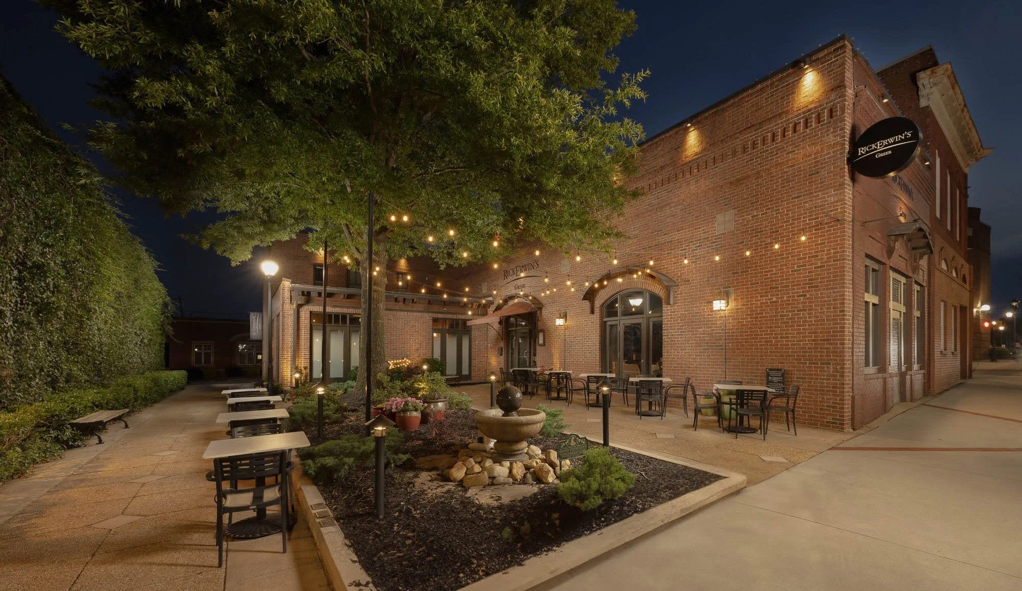Night view of a brick building with string lights, outdoor seating, green trees, and decorative plants.