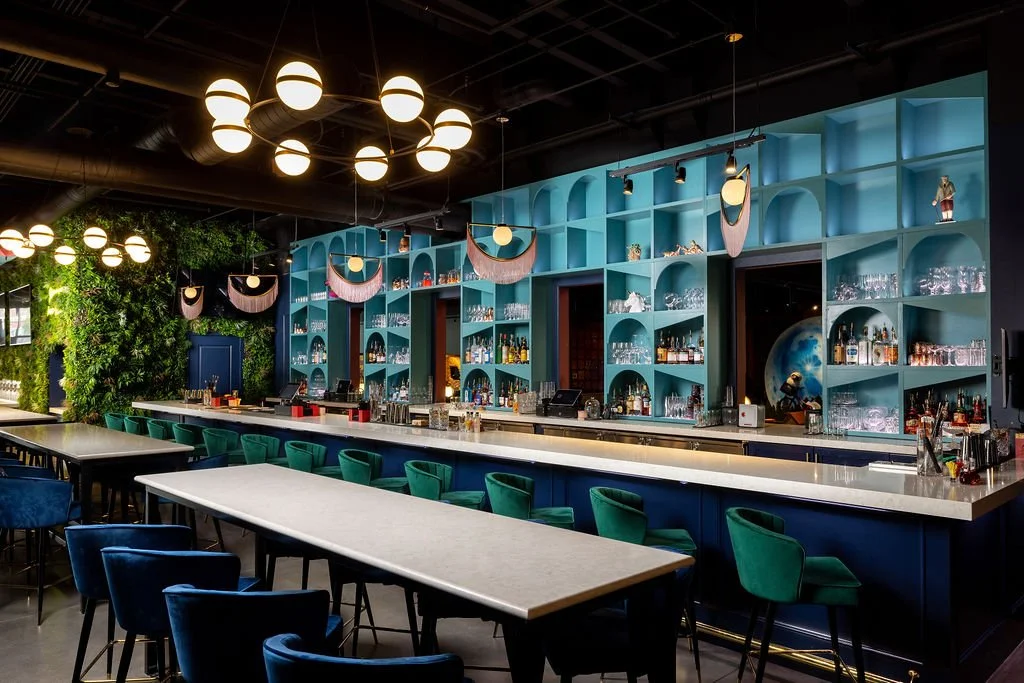 A stylish bar with a long white marble counter, blue and green chairs, a blue wall with built-in shelves displaying glassware and bottles, modern hanging light fixtures, and greenery on one side.