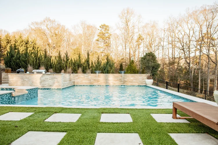 A backyard swimming pool with a stone fountain feature, surrounded by greenery and a wooden bench, with leafless trees in the background.