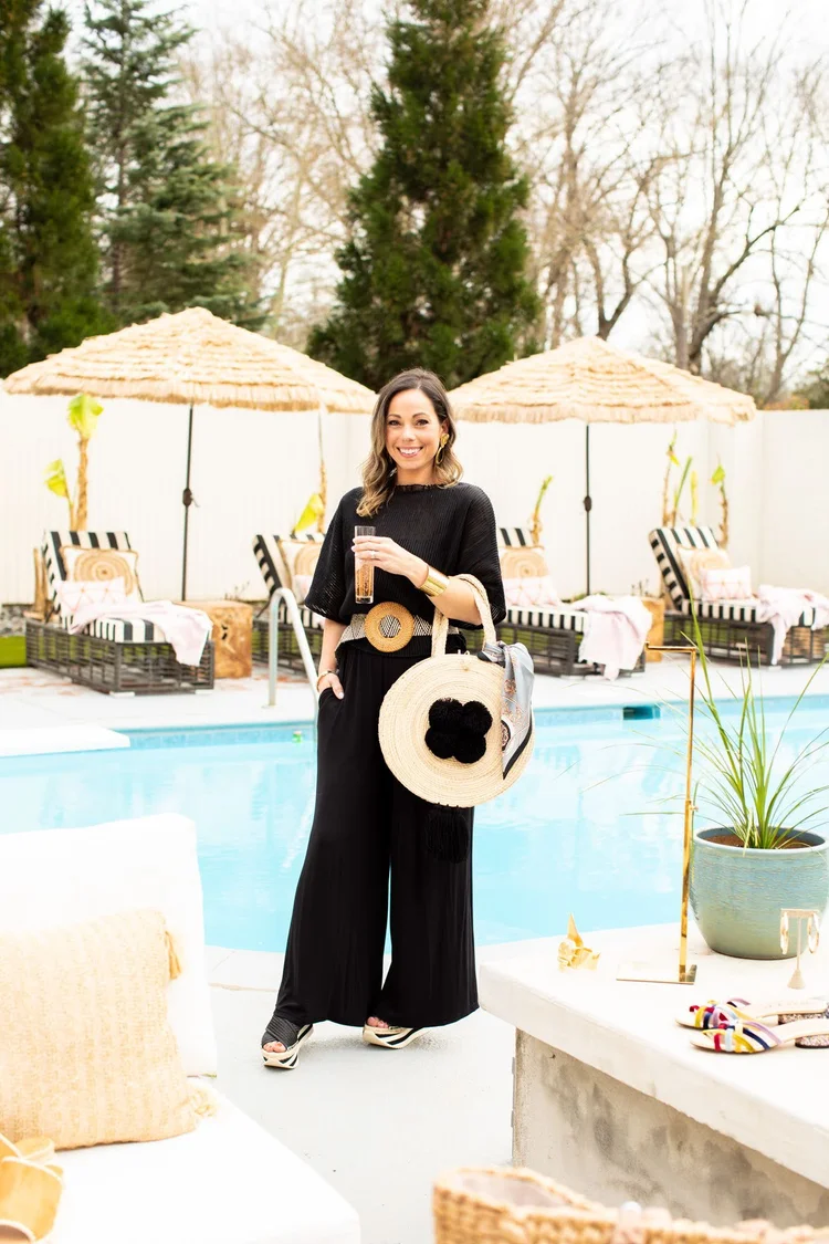 Woman in a black outfit holding a glass of drink by a poolside with straw umbrellas and seating in the background.
