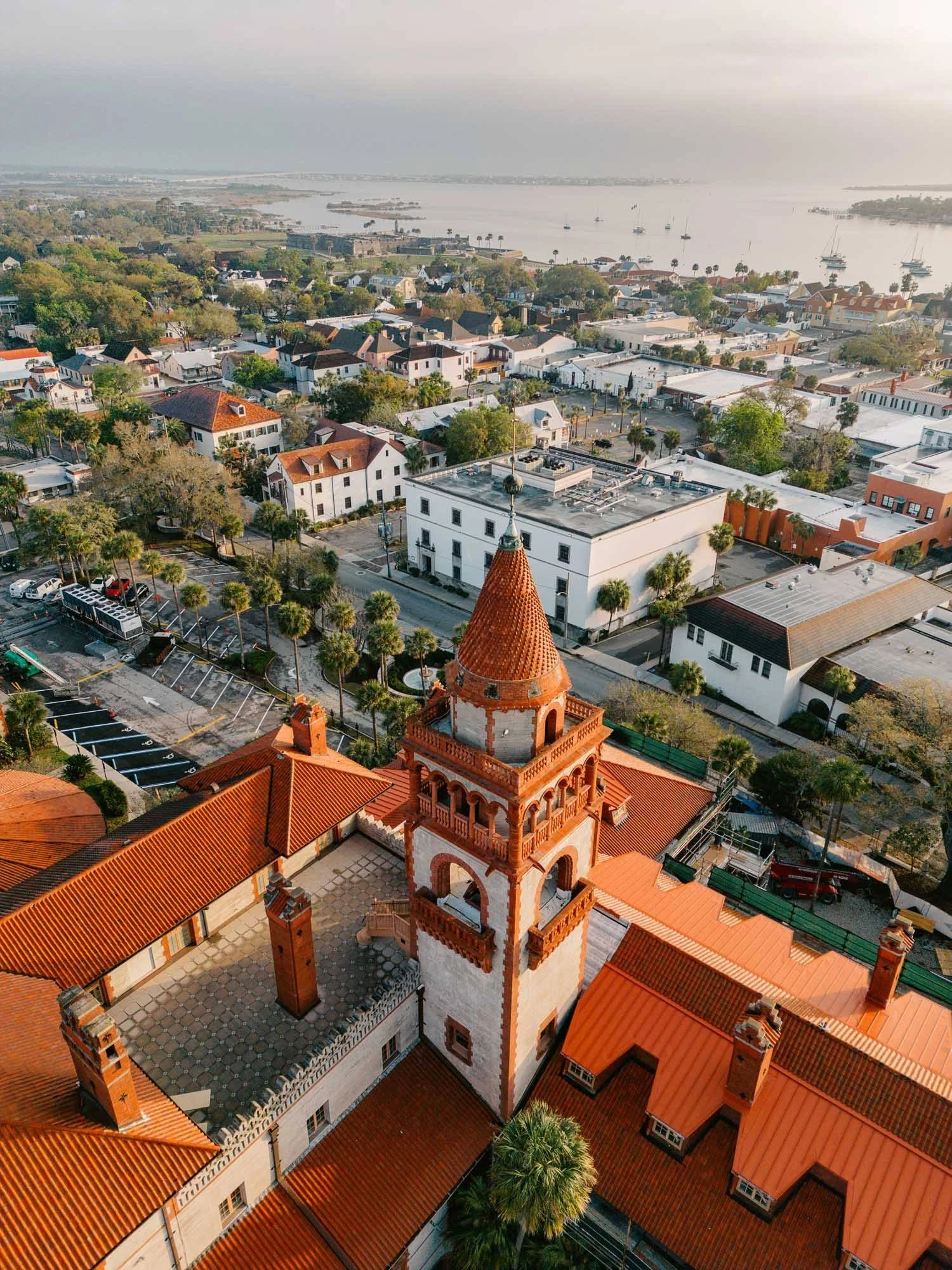 An aerial view of a coastal town featuring a red-tile rooftop church with a tall tower, surrounded by residential buildings and palm trees. In the background, there is a large body of water with sailboats and a shoreline dotted with more buildings an