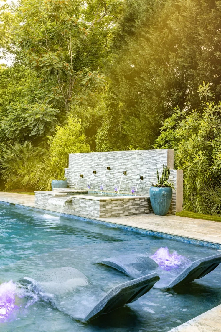 Swimming pool with built-in hot tub and waterfall feature, surrounded by lush green trees and plants in a backyard setting.