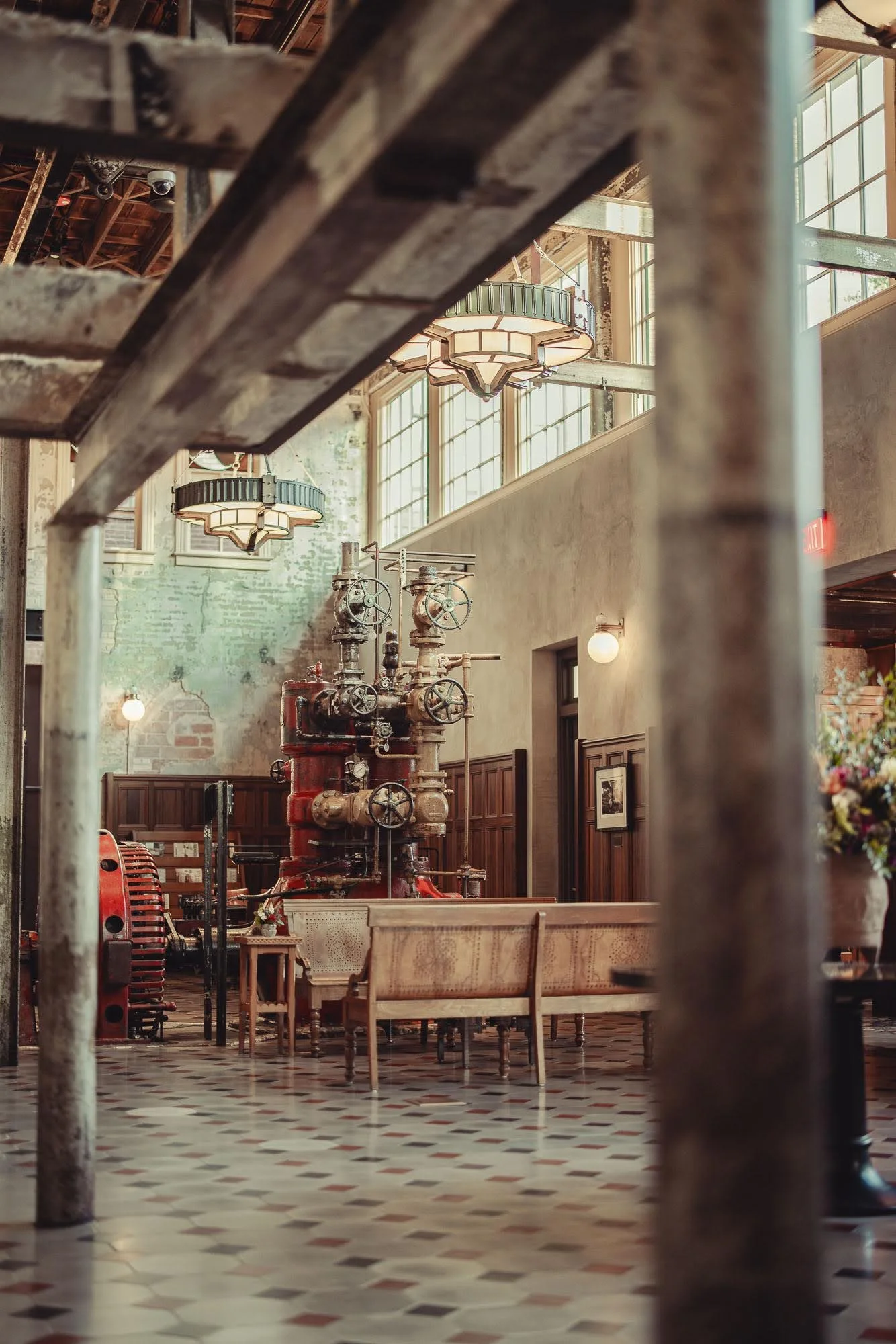 Interior of a vintage-style building with large windows, hanging light fixtures, industrial machinery, wooden benches, and a patterned tile floor, seen through metal bars.