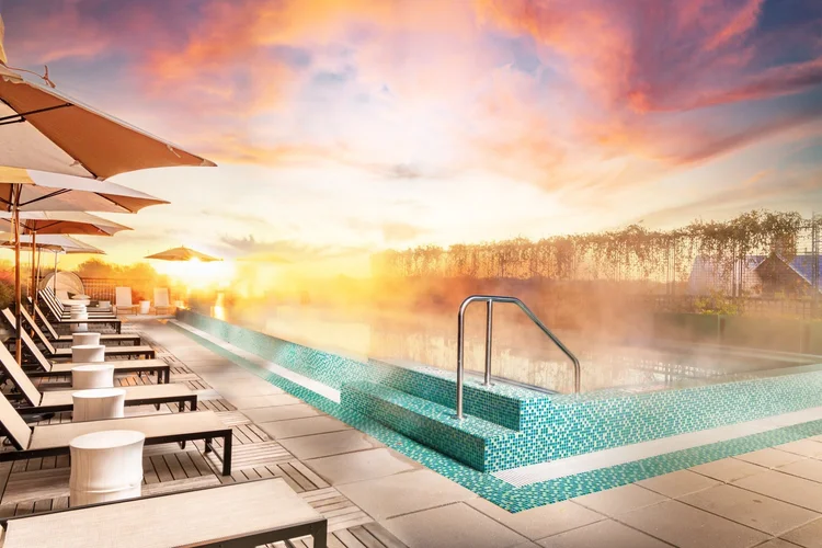 An outdoor pool area at sunrise with sun loungers, umbrellas, and steam rising from the hot water, under a colorful sky.