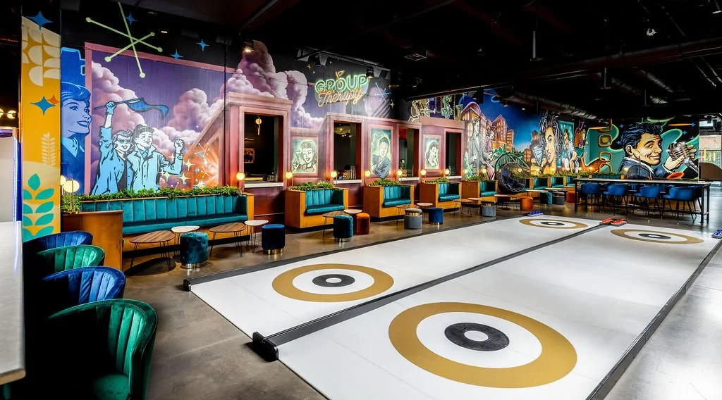 Indoor entertainment space with a shuffleboard court and colorful mural artwork on the walls, including people and abstract designs, with seating areas and tables.