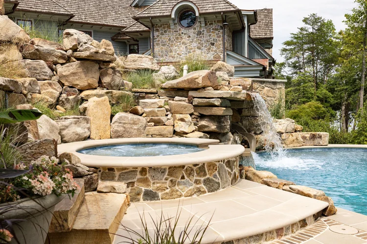 A backyard with a stone water slide leading into a pool, built on a rocky hillside with plants and trees around.