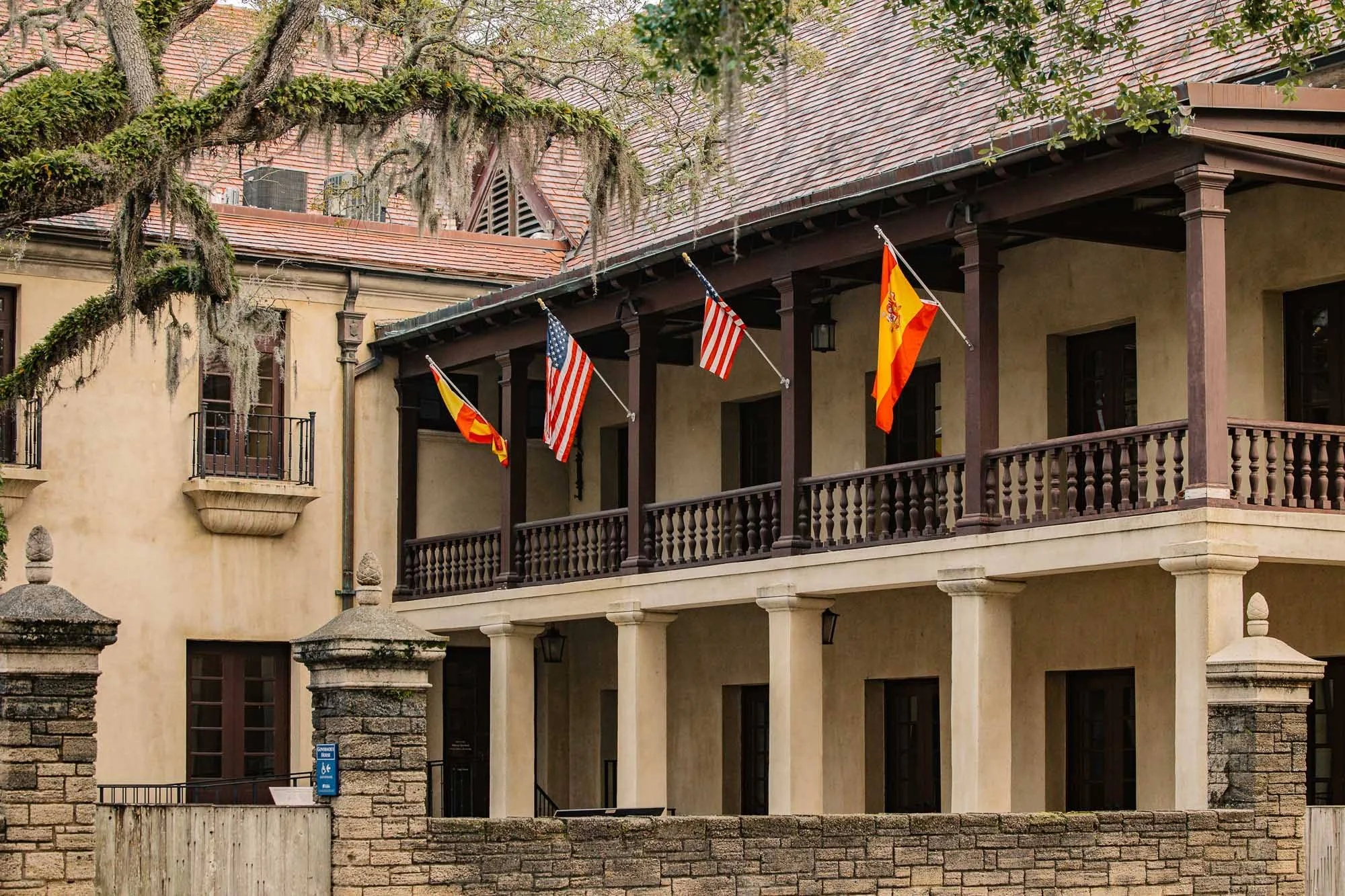 A building with a beige stucco exterior, stone accents on the lower level, and a upper balcony with multiple French doors. Flags, including the American, Spanish, and Catalan flags, are hanging from the balcony. There are large trees with moss hangin