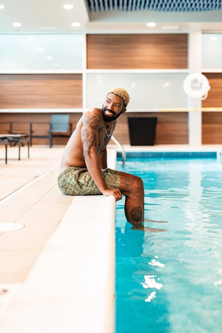 A smiling man with bleach blonde hair and tattoos sitting on the edge of an indoor swimming pool, with his feet in the water.