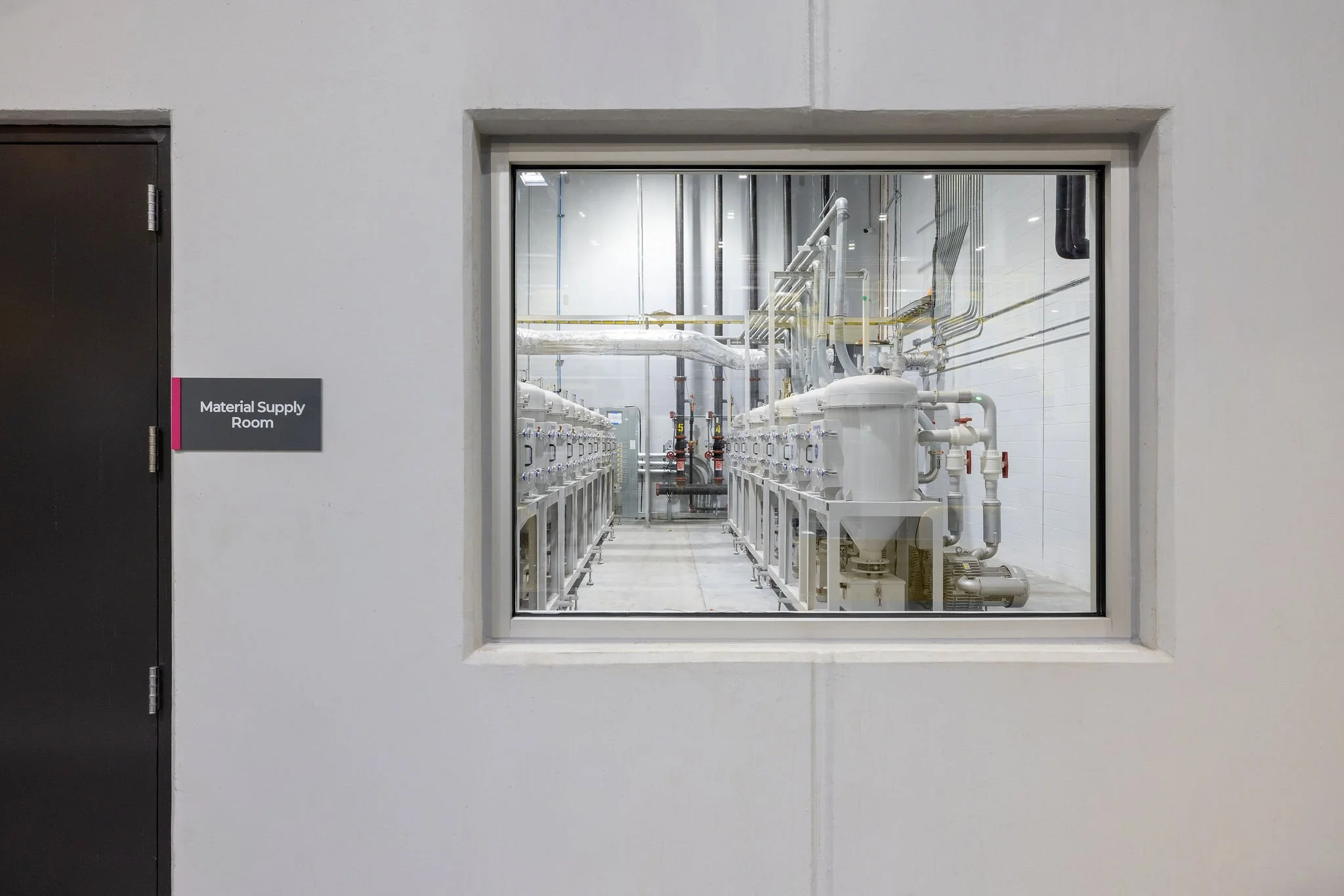 View through a glass window into a material supply room with white pipes and machinery.