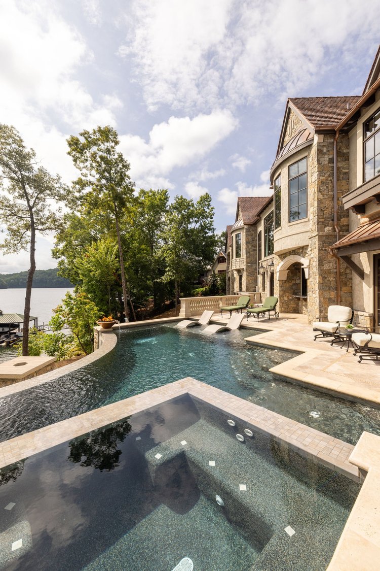 Luxury house with stone facade and large windows overlooking a swimming pool with a hot tub, near a waterfront, with trees and a partly cloudy sky in the background.