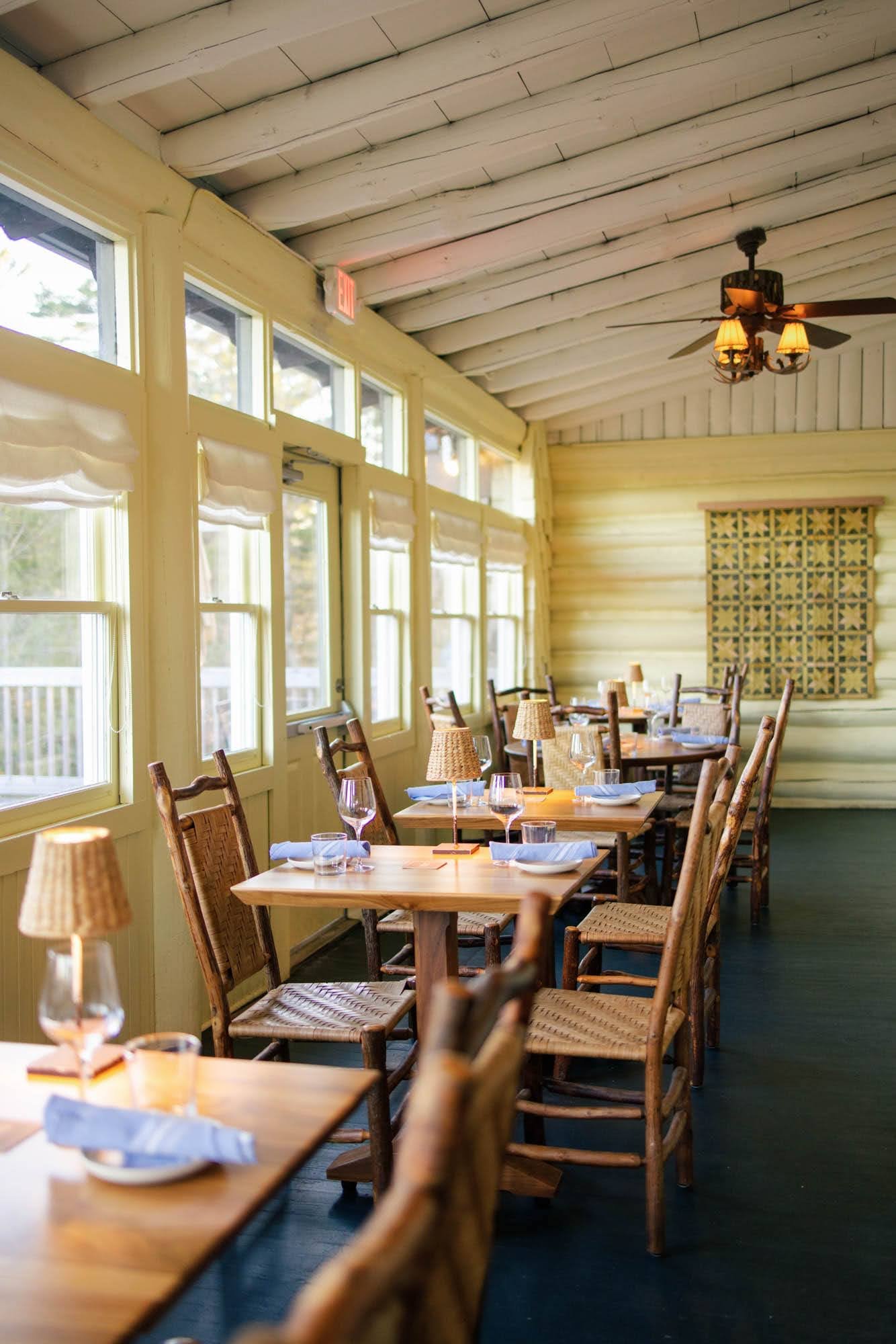 Inside view of a restaurant with wooden tables and chairs, set with wine glasses, napkins, and small lamps. Large windows line the wall, with sheer curtains, and a quilted wall hanging is visible in the background. The ceiling has wooden plank paneli