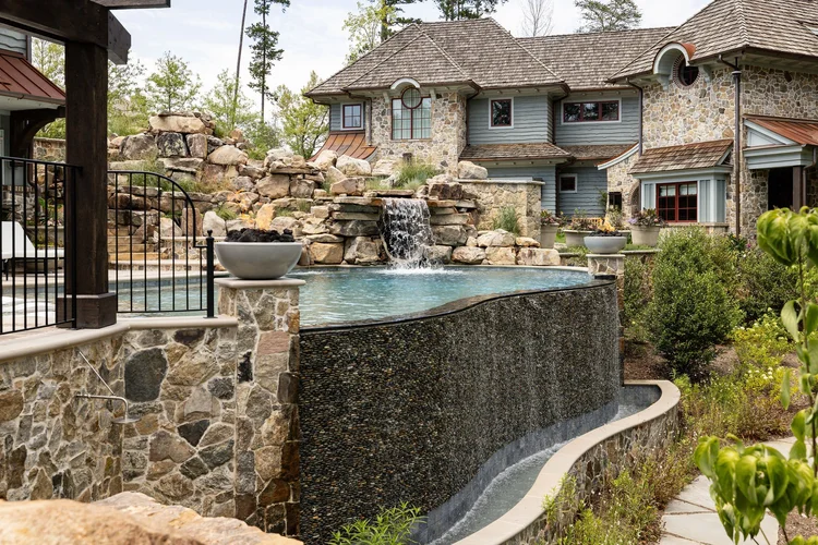 Luxurious backyard with a tiered waterfall, a swimming pool, stone and wood house, and lush greenery.