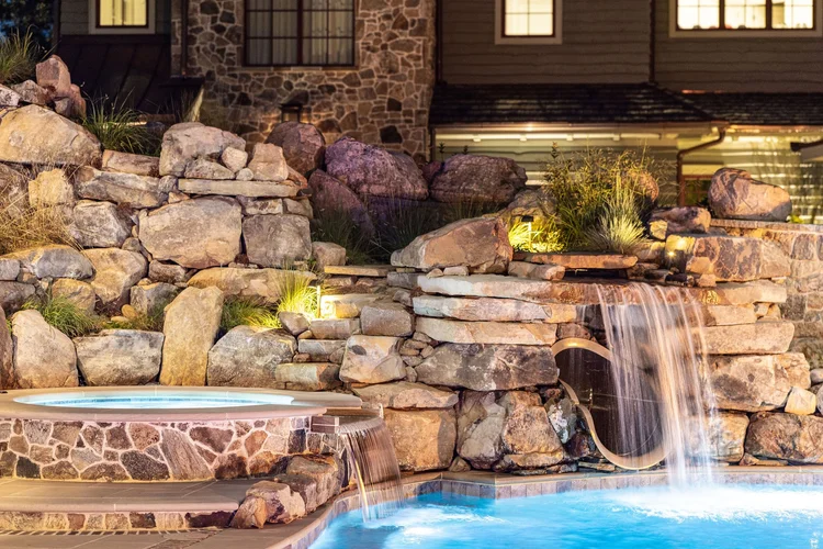 Nighttime view of a backyard with a stone waterfall feature flowing into a swimming pool, surrounded by rocks and plants with outdoor lighting.