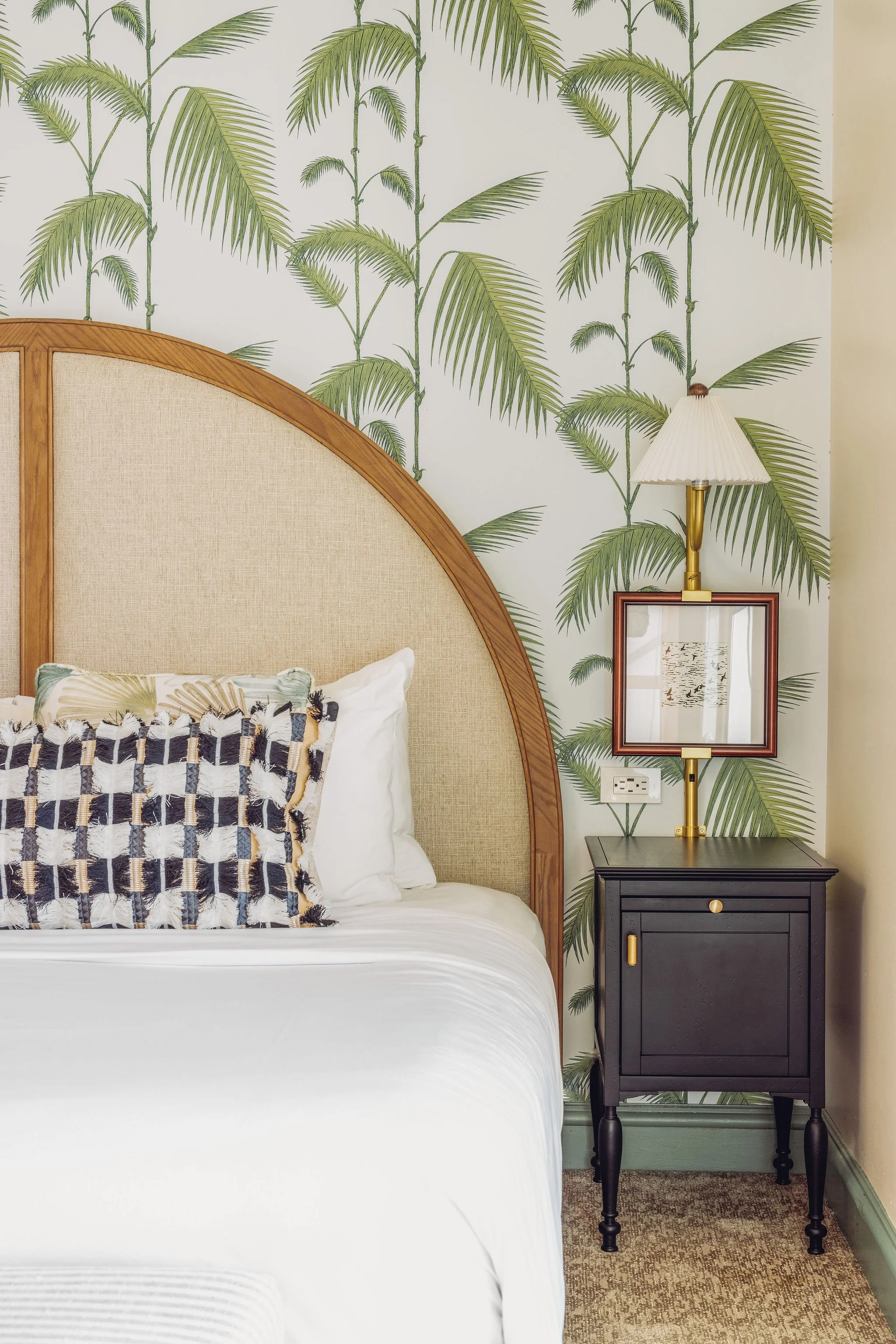 A bedroom with a bed, decorative pillow, and a black nightstand with a lamp and picture frame, against a wall with tropical palm leaf wallpaper.