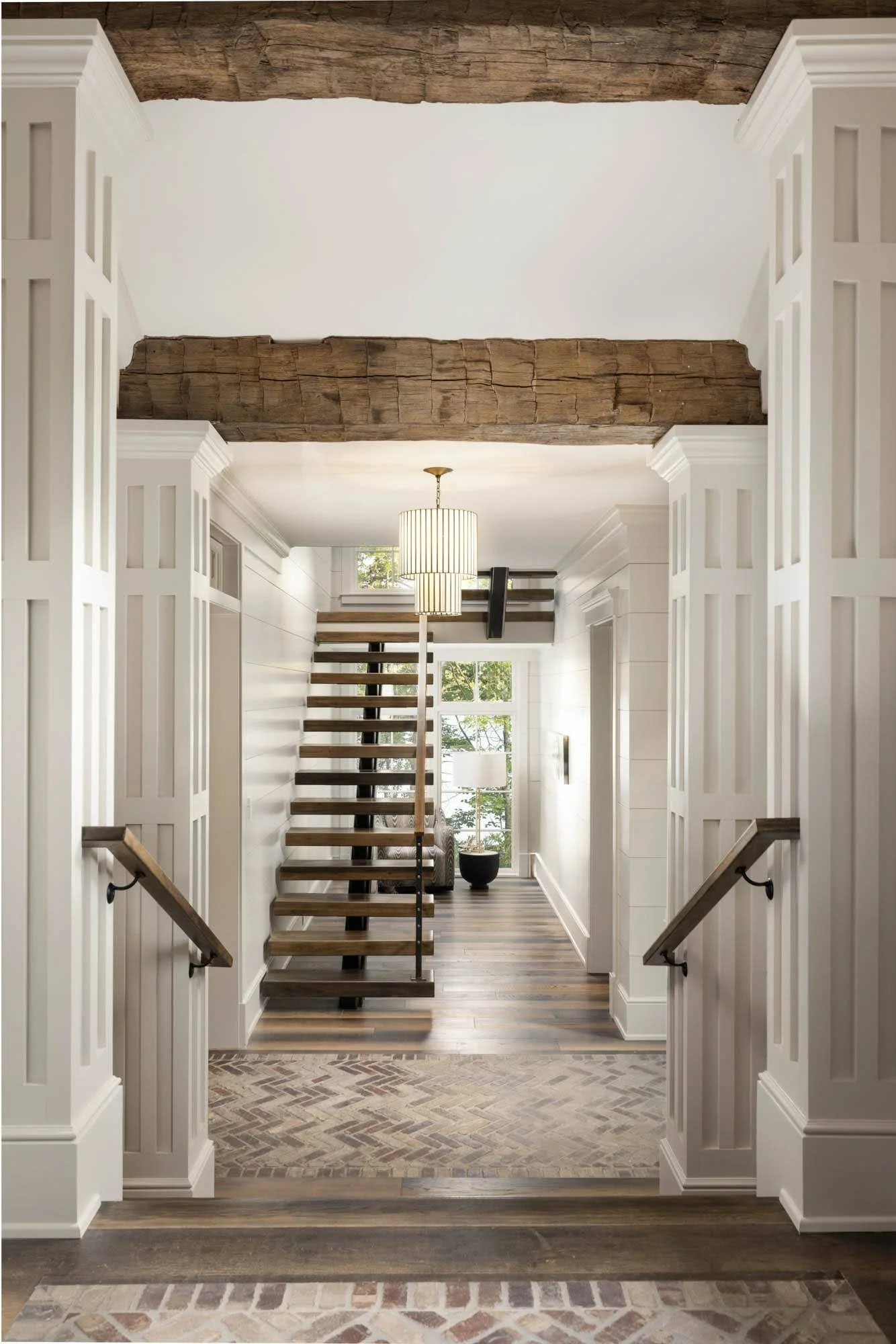 Interior view of a modern home with a hallway leading to a staircase and living area, featuring white walls, wooden accents, and large windows with greenery outside.