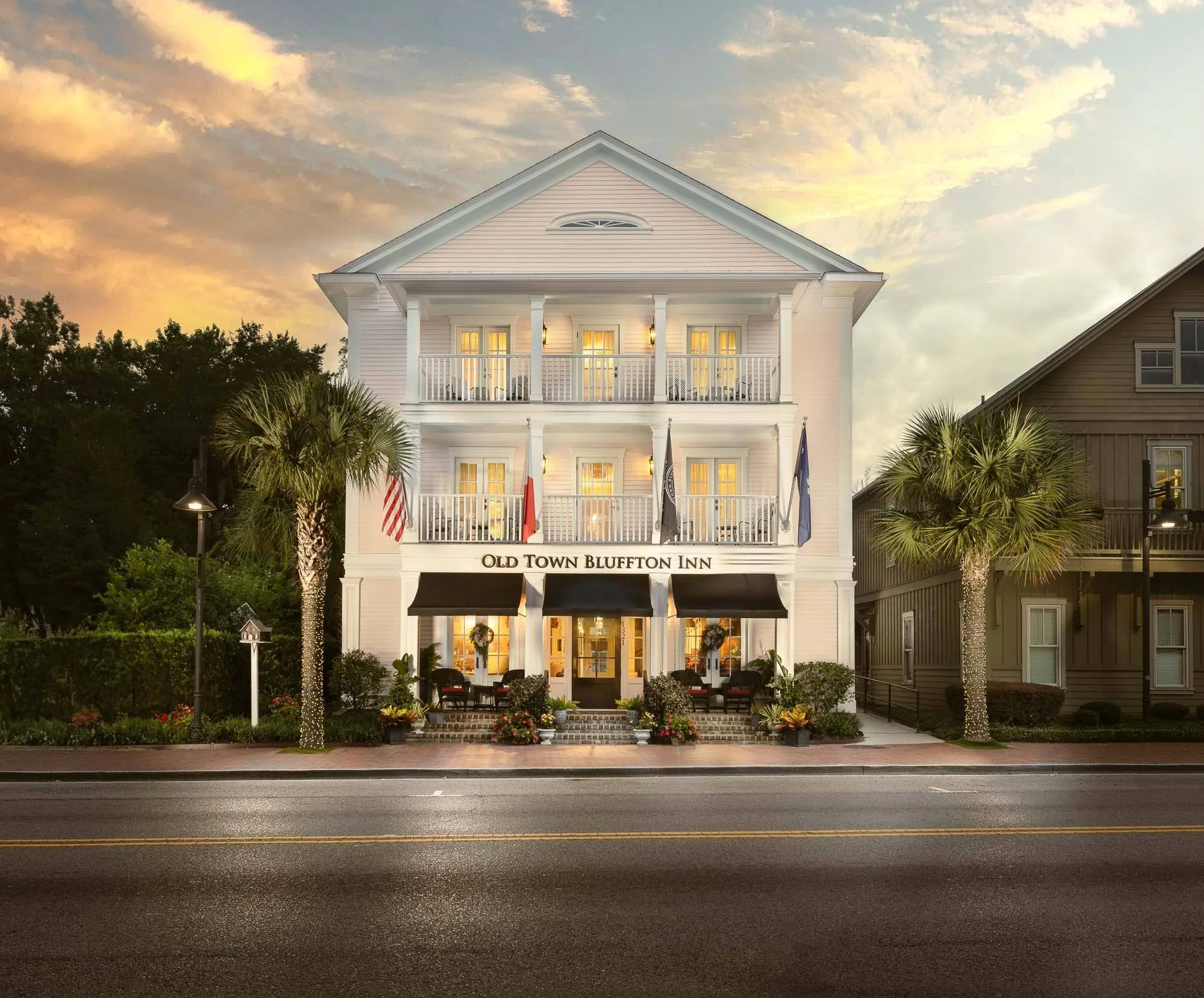 A white, three-story building labeled 'Old Town Bluffton Inn' with flags on the front porch, decorative lights on two palm trees, and outdoor seating, against a sky with clouds at sunset.