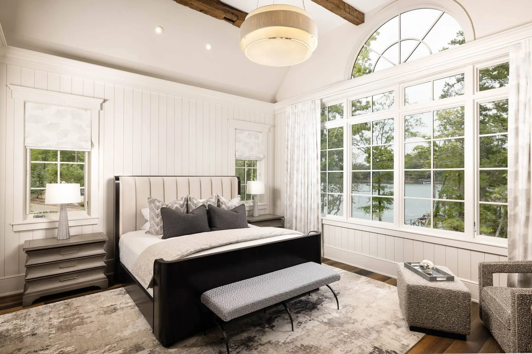 A bedroom with white walls, large windows showing a lake and trees outside, a bed with gray and black pillows, a bench at the foot of the bed, and a patterned rug.