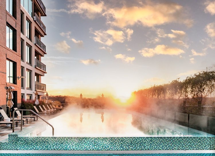 A rooftop swimming pool at sunset, with lounge chairs and umbrellas along the side, adjacent to a multi-story residential building.