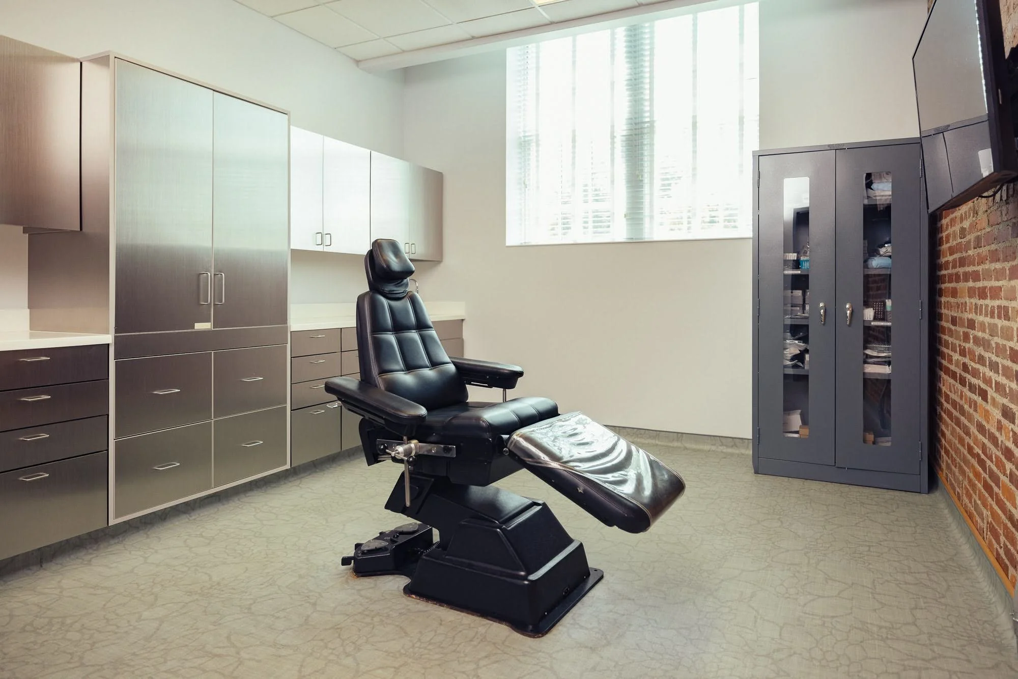 A medical examination room with a black reclining chair, white cabinets, storage cabinets with glass doors, and a brick accent wall, illuminated by natural light from large windows with blinds.