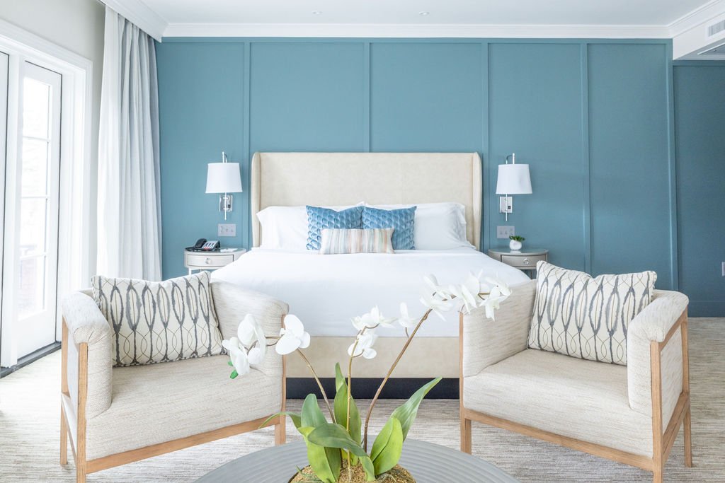 A bright hotel room with a blue accent wall, a white bed with a padded headboard, two nightstands with white lamps, a beige sofa with patterned pillows, white curtains, and a glass door leading outside. A potted plant with white flowers is in the for