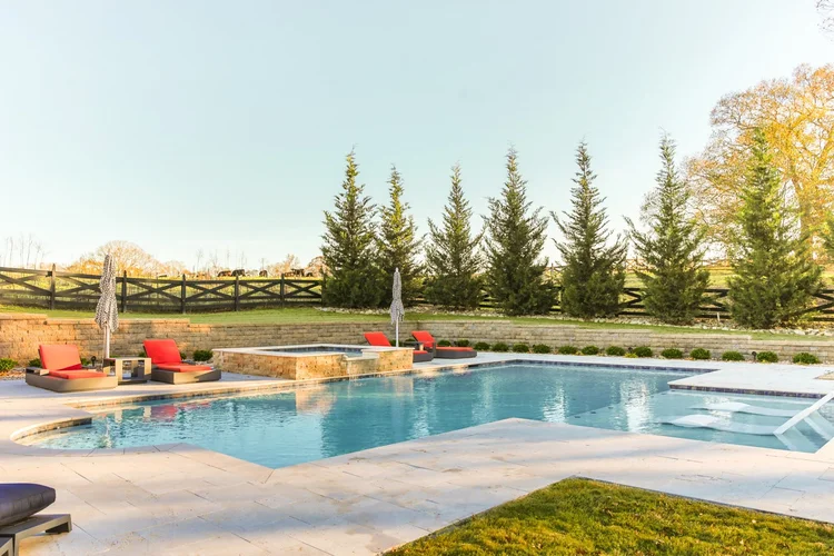 Outdoor swimming pool with lounge chairs and umbrellas, landscaped backyard with tall evergreen trees, stone retaining wall, and a grassy area, under a clear blue sky.