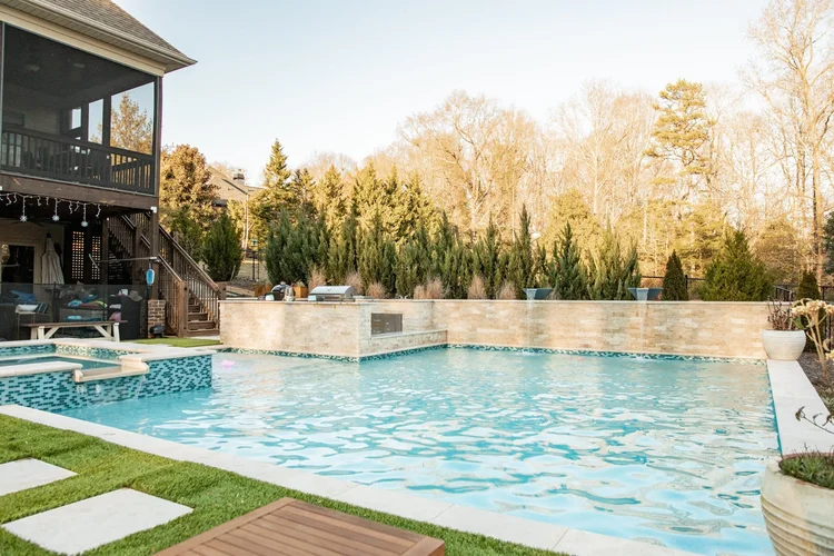 Backyard swimming pool with clear water, a beige stone wall with three waterfalls, and an outdoor kitchen area with grills, surrounded by lush green trees and a two-story house with a screened porch and outdoor seating, under a clear sky.