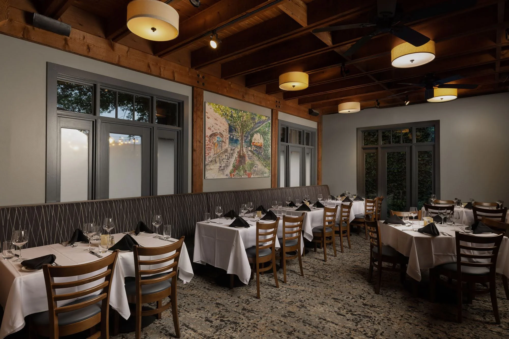 Dining room with tables covered in white tablecloths, black napkins, and glassware. Wooden chairs surround the tables. The room has large windows with black frames, a colorful painting on the wall, and a wooden ceiling with hanging lights and ceiling