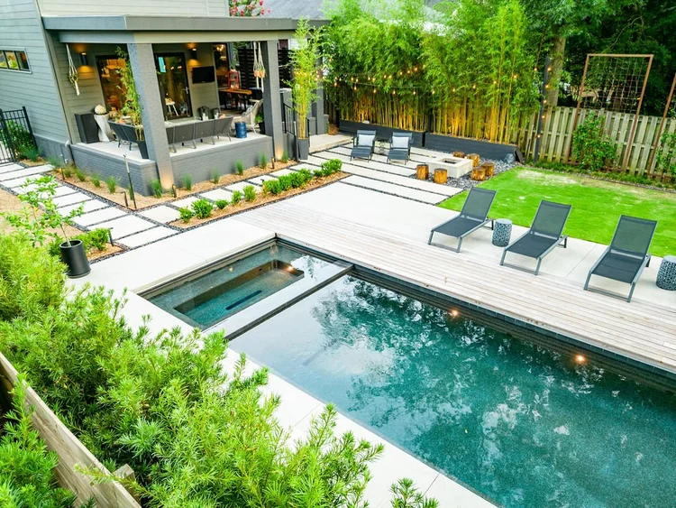 Backyard with a swimming pool, lounge chairs, and outdoor furniture, adjacent to a house with a covered patio and string lights, surrounded by greenery and trees.