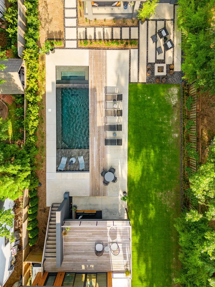 Aerial view of a backyard with a swimming pool, outdoor seating, a well-kept lawn, and surrounding greenery.
