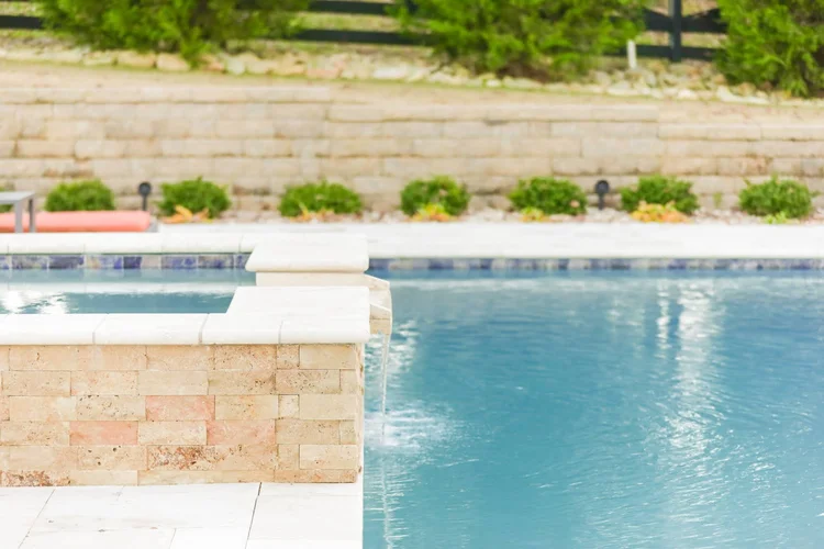 A swimming pool with a brick border and a relaxing backyard with greenery and a stone wall.